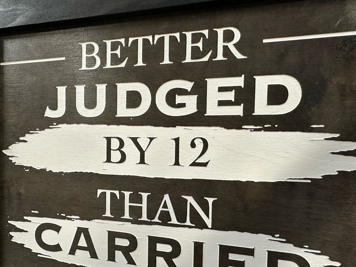 Dark brown wooden sign with white text reading “BETTER JUDGED BY 12 THAN CARRIED”.