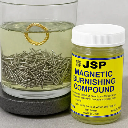Front view of JSP Magnetic Burnishing Compound jar beside a liquid tumbler with metal pins and a gold ring