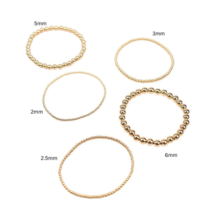 Five gold filled stretchy beaded bangle bracelets in 2, 2.5, 3, 5, and 6 millimeter bead sizes on white