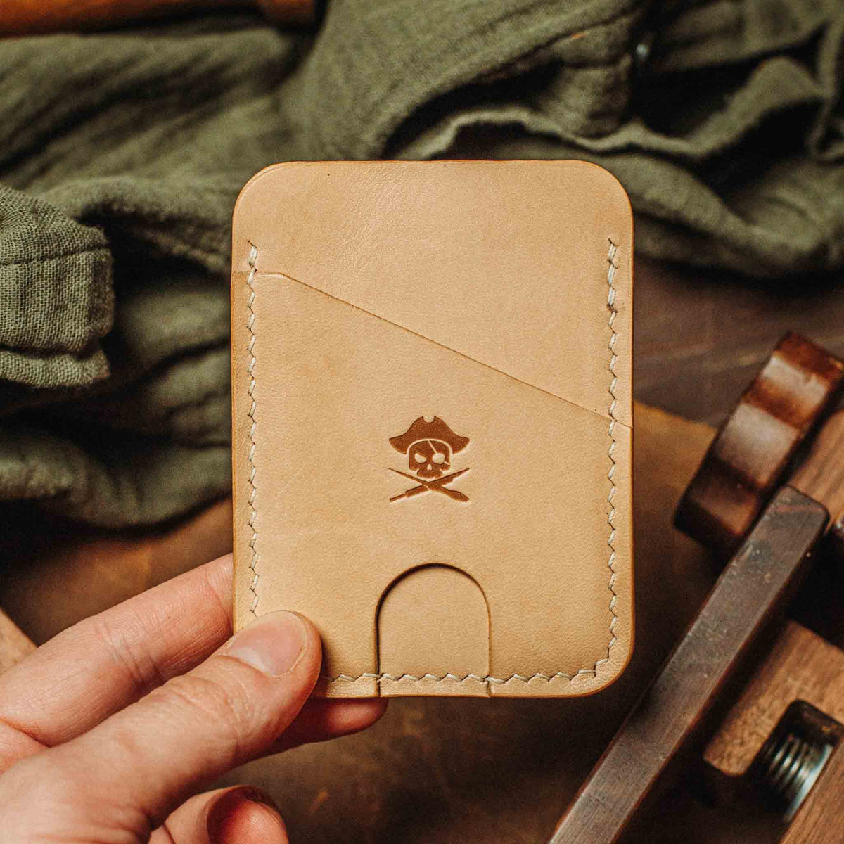 Tan leather Pirate Goods Boatswain v4 Slim Wallet with stitched edges and pirate skull emblem on a workshop bench