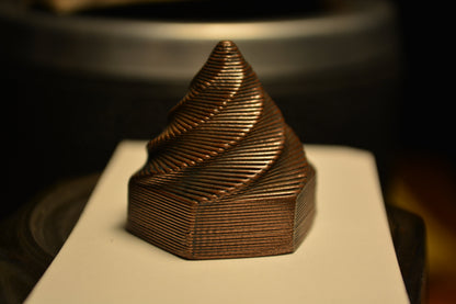 Copper-colored, 3D-printed metal test sample with a stepped, spiral pyramid shape on white paper.