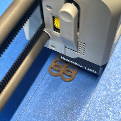A Bambu Lab 3D printer extrudes brown plastic, forming a connected 4-ring shape on a blue print bed.