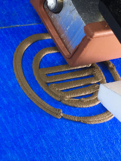 3D printer nozzle extruding gold filament in a circular coin shape on blue tape.