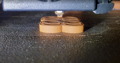 A 3D printer extrudes gold-colored filament to form a textured, four-lobed plastic block on a textured bed.