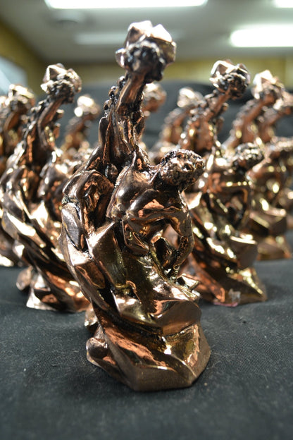 Copper-colored metallic statues of a figure lifting a rock, arranged on a dark surface.
