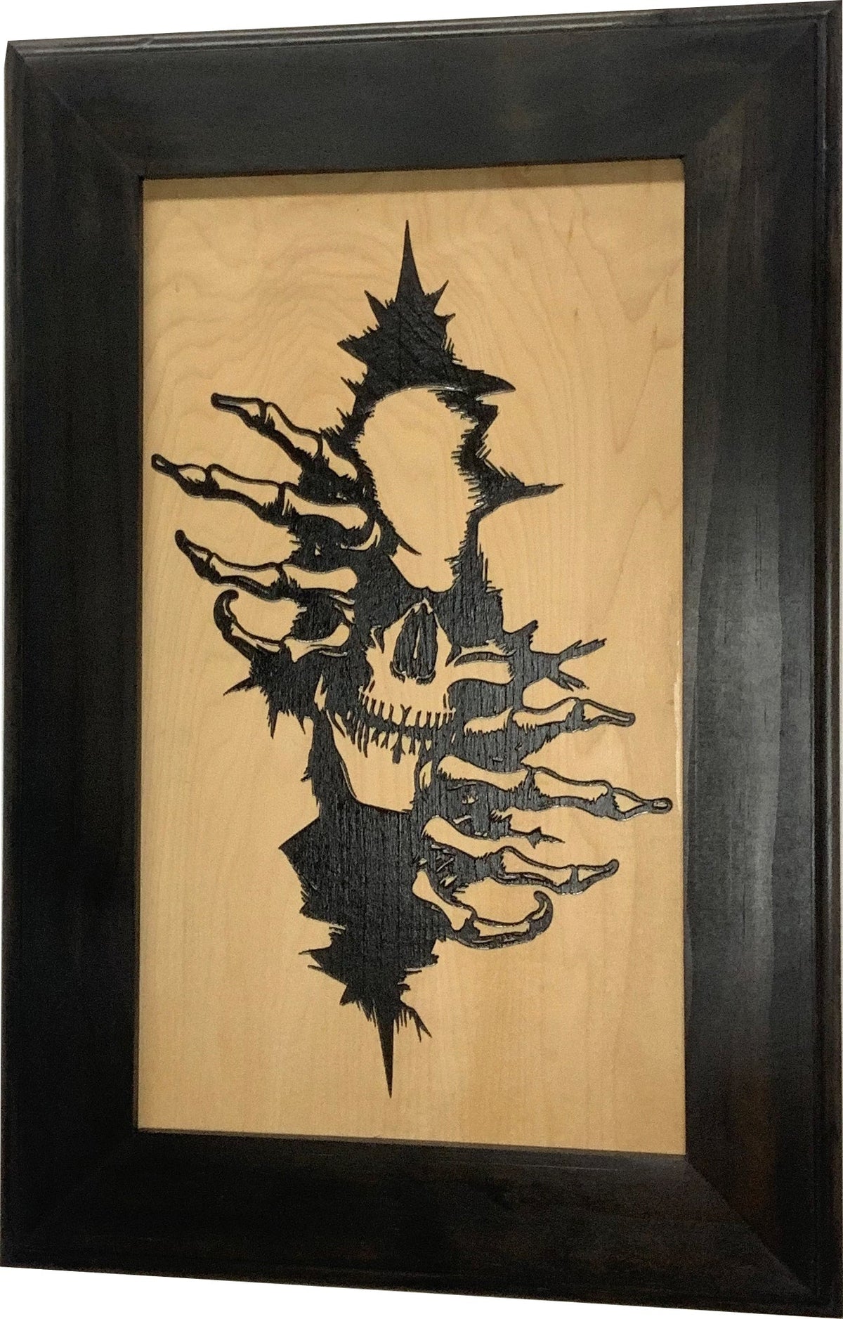 Framed wood-burned artwork of a skull and skeleton hands emerging from a jagged tear in light wood.