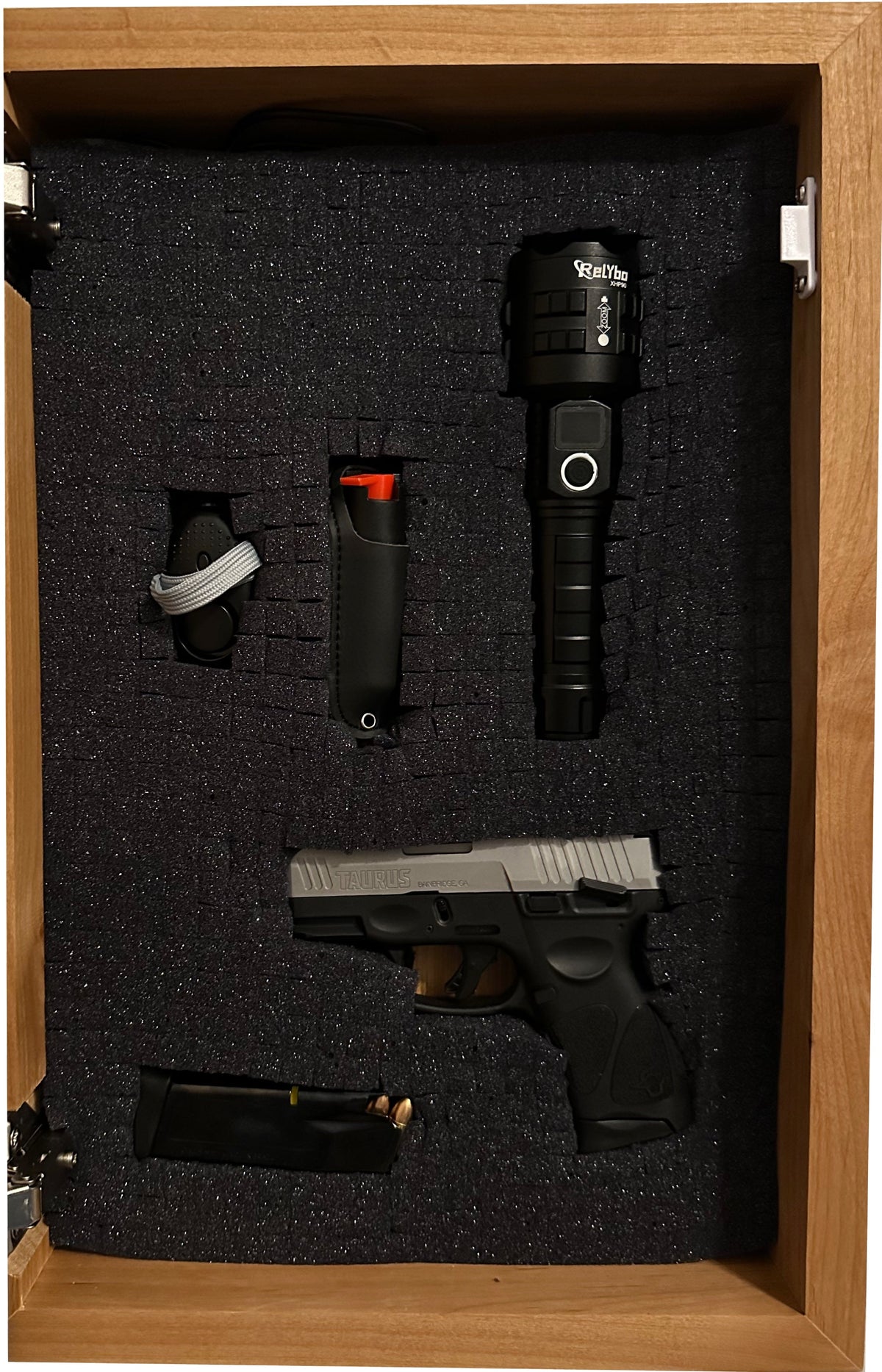 Open wooden cabinet with charcoal pluck foam interior holding a black-and-silver Taurus handgun, magazine, flashlight, and holster.