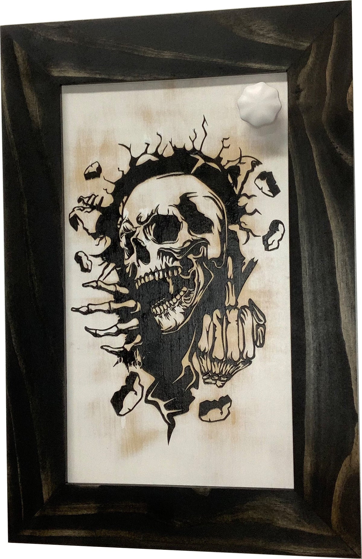 Wood-burned sign of a skull breaking through a wall, framed in dark wood, with a white knob.