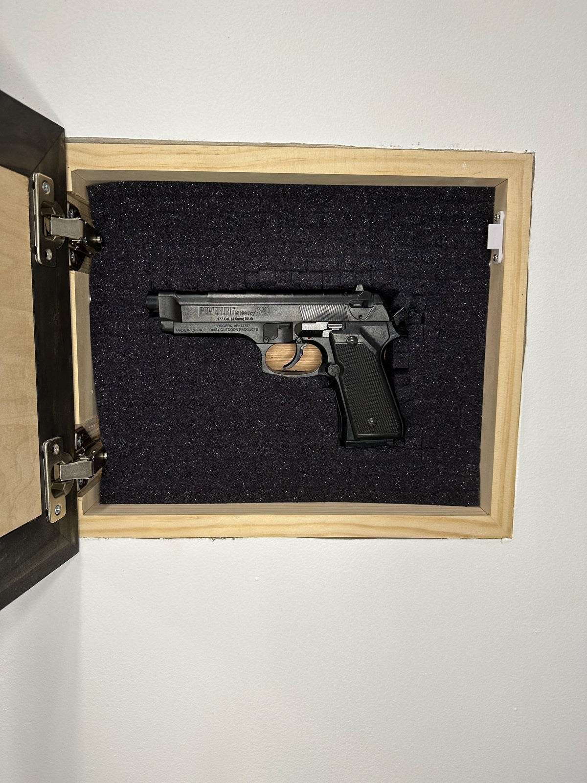 Black handgun securely stored in a wall-mounted wooden case with black protective foam interior.