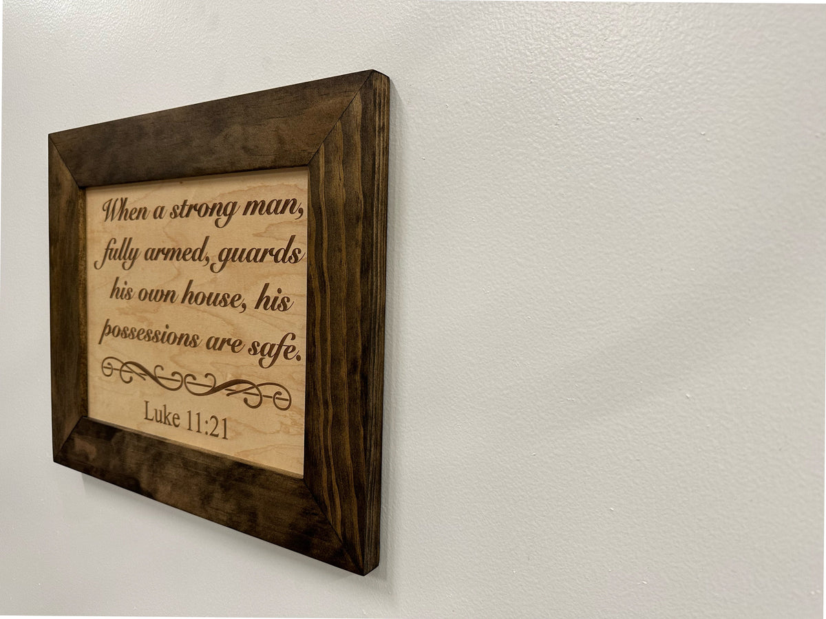 Engraved wooden sign with Luke 11:21 quote, in a dark brown wood frame on a wall.